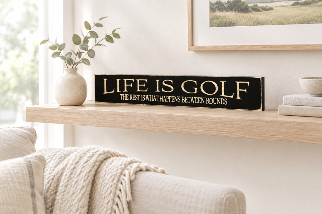 Black wooden sign with engraved lettering that reads "Life is Golf. The rest is what happens between rounds."  This sits on a natural wood shelf along with a plant books and a dish, in a cozy room setting.