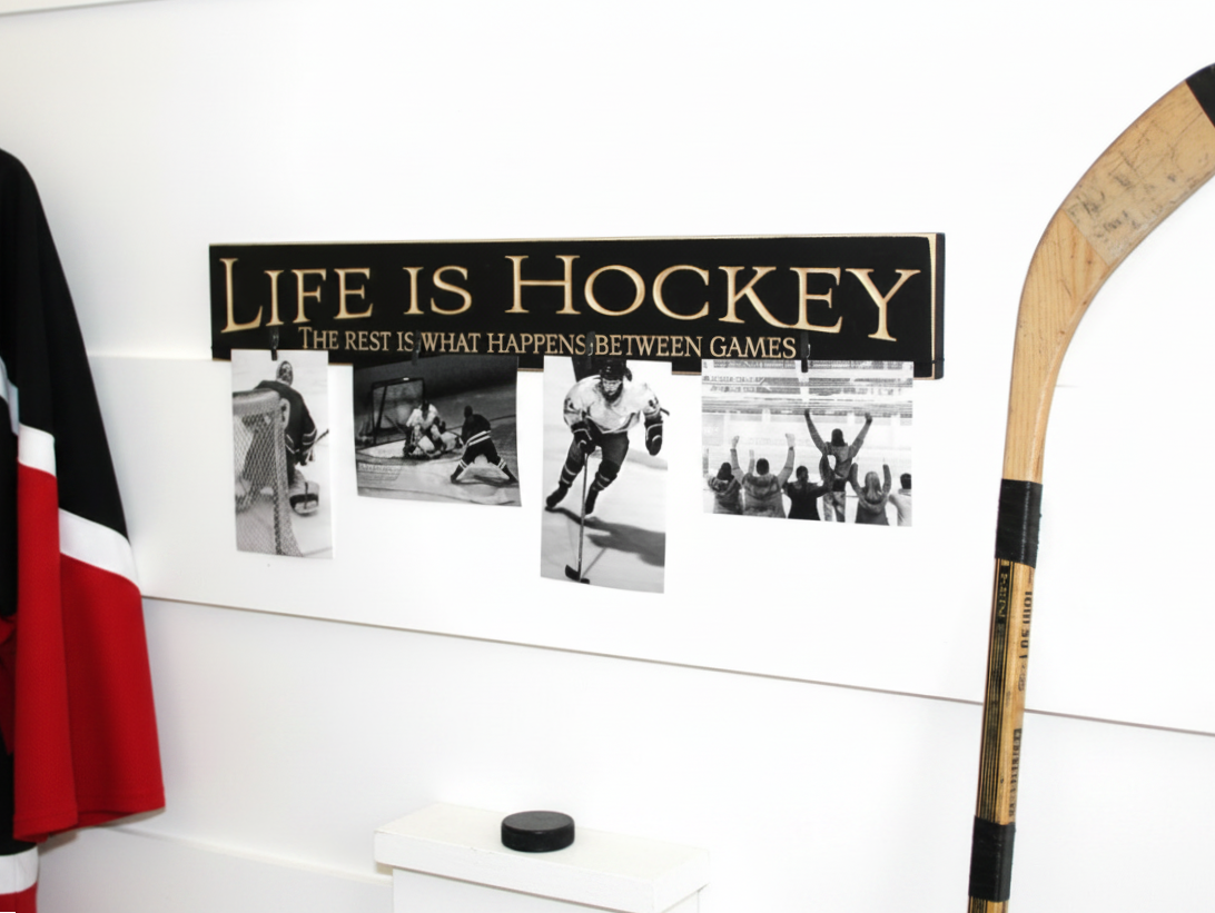 A hockey-themed wall sign with the text 'LIFE IS HOCKEY' and three black-and-white photos of players, displayed on a white wall with a hockey stick and jersey partially visible.