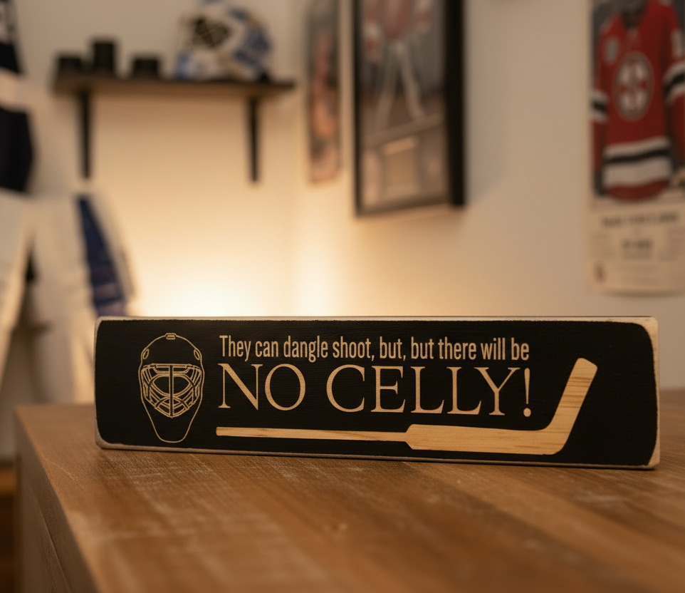 A rectangular black wooden hockey-themed sign with engraved lettering that reads 'They can dangle shoot, but, but there will be NO CELLY!' sits on a wooden table in a warmly lit room. The sign features a gold goalie mask graphic and a hockey stick illustration.