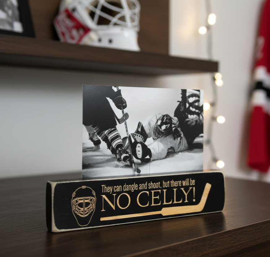 A black wooden hockey-themed photo stand with engraved lettering that reads 'They can dangle and shoot, but there will be NO CELLY!' and a goalie mask graphic, holding a black-and-white hockey action photo on a dark wooden shelf.