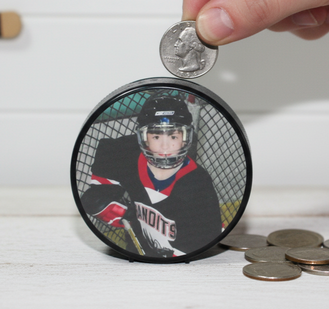 A hockey puck bank with a printed photo of a hockey player. A hand is placing a US quarter into the slot on top of the box, with other coins scattered nearby on a white wooden surface.