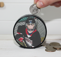 A hockey puck bank with a printed photo of a hockey player. A hand is placing a US quarter into the slot on top of the box, with other coins scattered nearby on a white wooden surface.