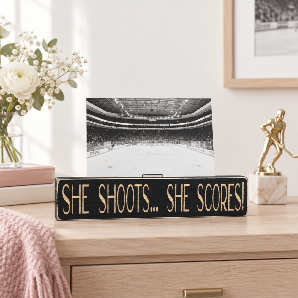 A black rectangular decorative sign with engraved lettering reading "SHE SHOOTS... SHE SCORES!"  This sits on a natural wood dresser with books, a vase with flowers, a trophy and a blanket in a cozy room setting with sunlight pouring in the window.
