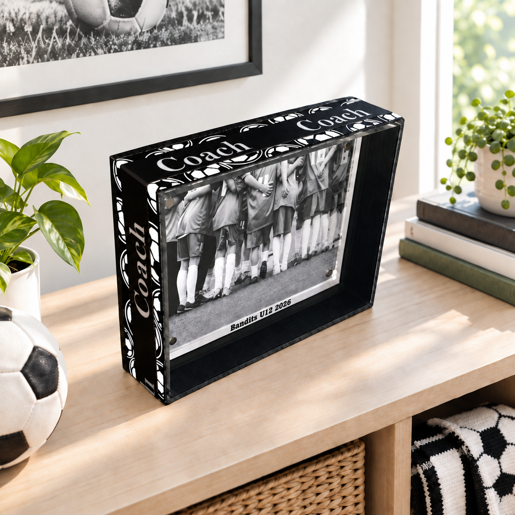 Black and white photo frame with a soccer ball pattern and COACH on the edges. The frame holds a black and white photo of a soccer game. The text "Bandits U12 2020" are visible just under the photo.  This sits on a natural wood table along with a plant and some books and a soccer ball in the background.