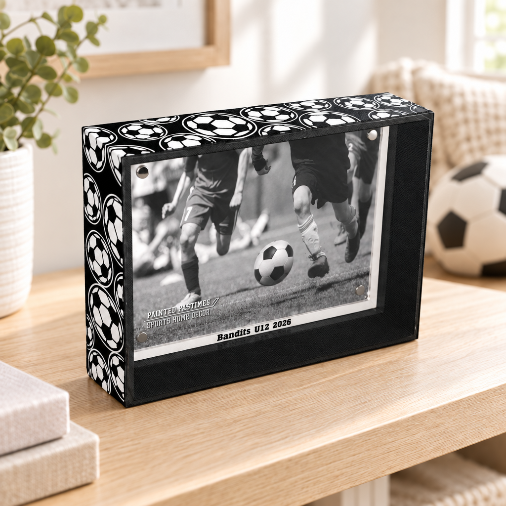 Black and white photo frame with a soccer ball pattern on the edges. The frame holds a black and white photo of a soccer game. The text "Bandits U12 2020" are visible just under the photo.  This sits on a natural wood table along with a plant and some books and a soccer ball in the background.