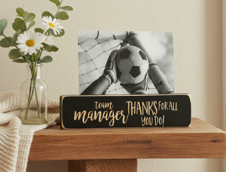 A rectangular black wooden sign with lasered lettering that reads 'team manager THANKS FOR ALL YOU DO!' sits on a wooden table next to a glass vase with white daisies. A black-and-white photo of soccer hands holding a ball is propped behind the sign.