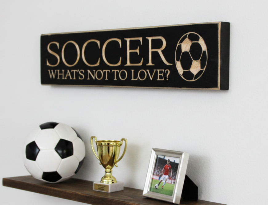 A rustic black wooden wall sign with engraved lettering that reads 'SOCCER WHAT'S NOT TO LOVE?' and a soccer ball graphic, displayed above a shelf with a soccer ball, gold trophy, and framed photo.