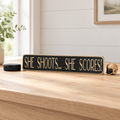 A rectangular black decorative sign with engraved lettering that reads “SHE SHOOTS... SHE SCORES!".   This sits on a natural wood table along with a puck, helmet and plant, in a cozy room with light pouring through.
