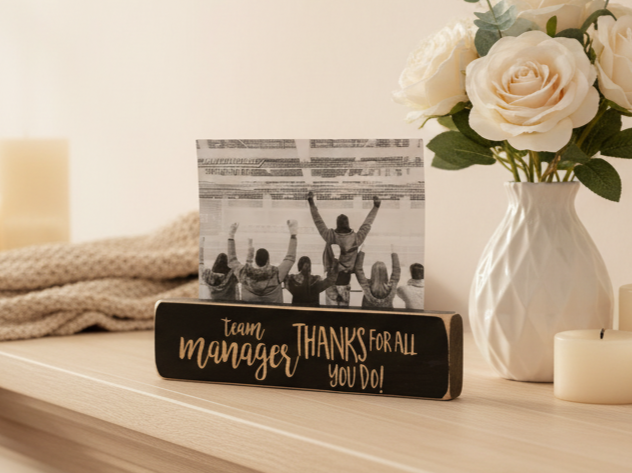 A personalized wooden photo block sign on a light wood table, styled with a black-and-white photo of a cheering crowd and lasered lettering that reads 'team manager THANKS FOR ALL YOU DO!'. White roses in a textured vase and candles are in the background.