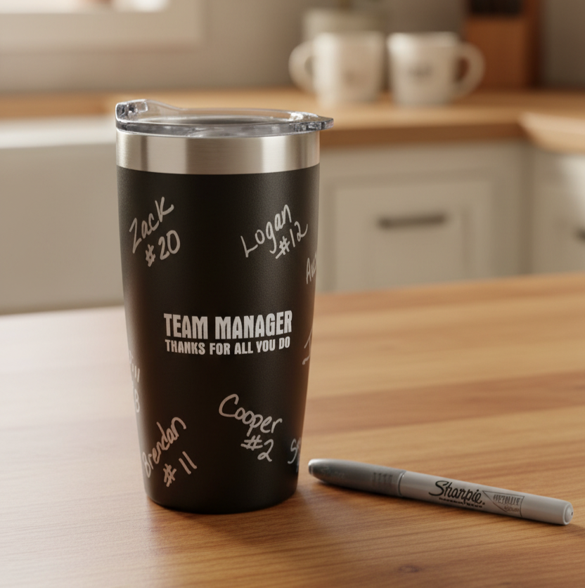 A black insulated tumbler with a clear lid sits on a wooden table in a kitchen setting. The tumbler has white handwritten signatures and the text 'TEAM MANAGER THANKS FOR ALL YOU DO'.  This comes with a free marker for the team to sign.