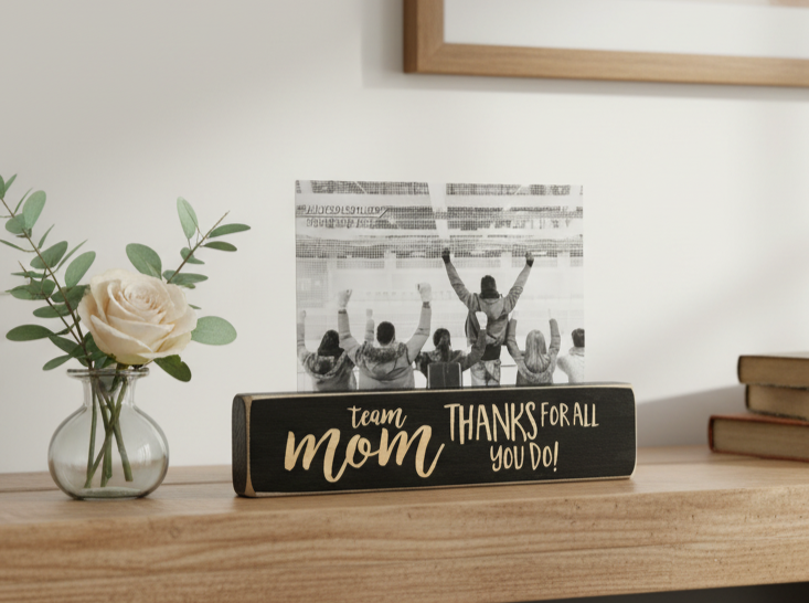 A decorative black wooden sign with lasered lettering that reads 'team mom THANKS FOR ALL YOU DO!' sits on a wooden shelf beside a small glass vase with a white rose and stacked books. A black-and-white photo of a crowd is displayed behind the sign.