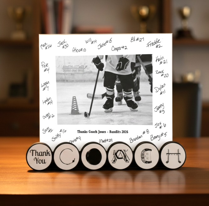 A custom hockey-themed photo frame display on a wooden table. The white rectangular frame is personalized with handwritten names around a black-and-white photo of ice hockey players, with 'Thanks Coach Jones - Bandits 2016' written at the bottom. This is displayed in hockey pucks the spell out COACH in unique hockey letters and Thank you.
