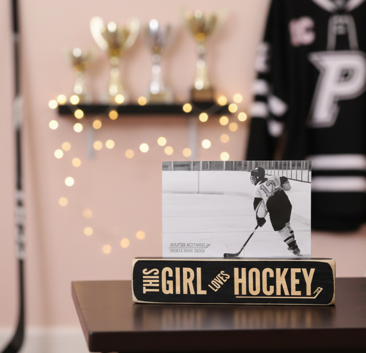 A custom hockey-themed wooden block sign on a dark table, featuring a black and white photo of a hockey player with the engraved text 'THIS GIRL LOVES HOCKEY'. The background shows a cozy room setting with a pink wall, string lights, trophies, and a hockey jersey.