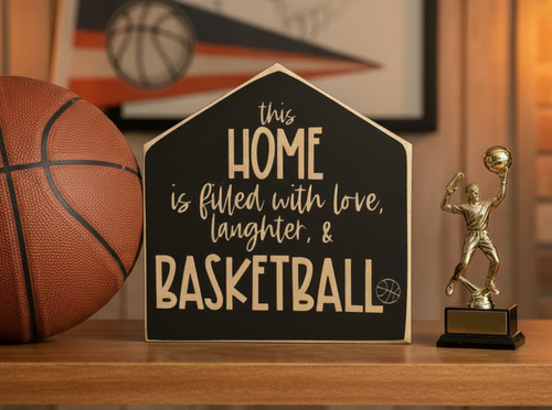 A house-shaped black wooden sign with engraved lettering that reads 'this HOME is filled with love, laughter, & BASKETBALL' sits on a wooden shelf next to a basketball and a small gold basketball player trophy.