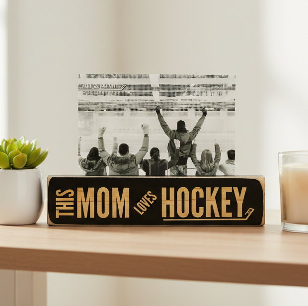 A rectangular wooden sign on a light wood table reads “THIS MOM LOVES HOCKEY” in engraved lettering on a black background. Behind the sign, a black-and-white photo of a hockey crowd with raised arms is displayed.