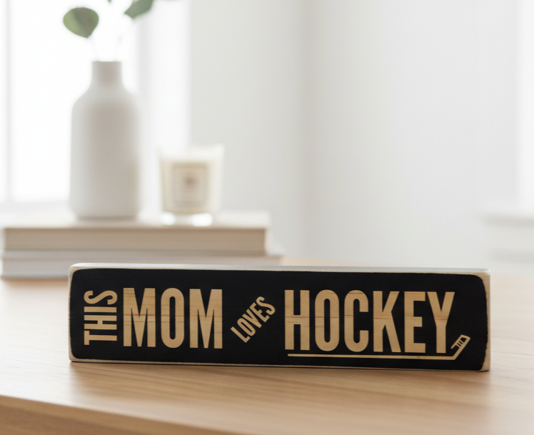 A rectangular black wooden sign with engraved lettering that reads 'THIS MOM LOVES HOCKEY' sits on a light wood table in a softly blurred indoor setting with books and a white vase in the background.