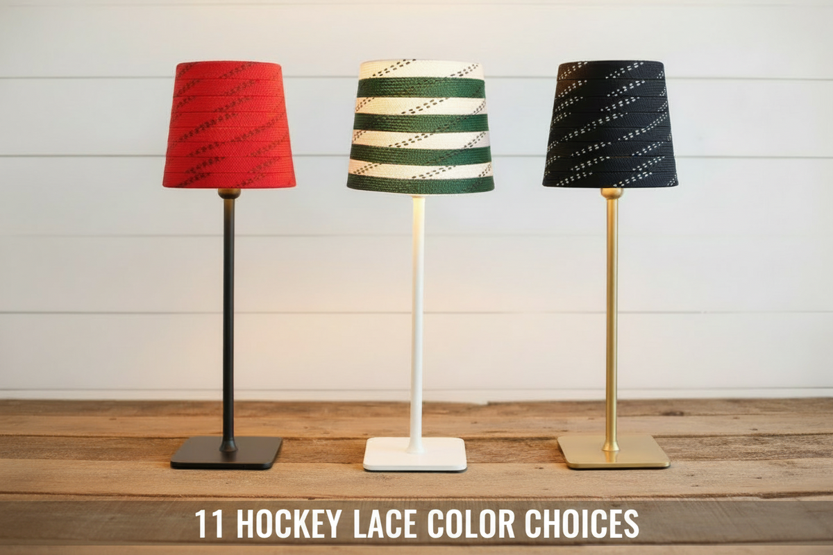 Three table lamps with hockey lace  shades stand on a wooden surface against a white shiplap wall. The left lamp has a red hockey lace shade and a black stand; the center lamp has white and green hockey lace shade and a white stand; the right lamp has a black hockey lace shade with a brass stand.